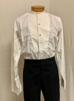 "1900, 15 1/4\" Neck, white cotton collarless shirt with pique bib front, 3 button holes for studs.  Shirt opens to hem in front, and buttons down at back 3/4 way. Curved at each hip at bottom.  Sleeves are long with  double French cuffs. Label reads \" Simpson Piccadilly. Measurements: neck 15 1/4\" Chest 42\" Shoulder to shoulder 16\" Shoulder to hem 30\" Sleeves 25\" Arm holes 9\" Width at bottom 44\"" Classic Cotton Shirt With Stand Collar, Classic Shirt With Stand Collar And Buttons, Classic White Shirt With Stand Collar, White Shirt With Covered Buttons For Daywear, White Dress Shirt For Daywear, Classic Shirt With Stand Collar And Placket, White Formal Shirt With Stand Collar, Formal White Shirt With Stand Collar, Collarless Shirt