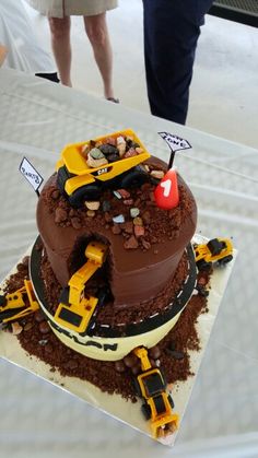 a chocolate cake with construction vehicles on it and people looking at the cake in the background