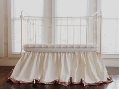 a white crib with pink ruffled bedding in front of two windows and wooden floors