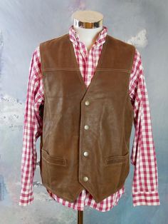 This 1990s brown leather vest has a V neckline, and closes in the front with four snap buttons. The vintage pointed-front Western waistcoat has two pockets on the front, and features topstitching on the leather. The vest has a full beige canvas (cotton) back with a leather half-belt with buckle. Chest = Up to 48 inches (121.92cm) Vest Length (front) = 26 inches (66.04cm) Vest Length (back) = 24 inches (60.96cm) Brand label: Pionier Sportive Size: 46 to 48 US/UK Material: Genuine Leather --- COND Brown Workwear Vest With Buttons, Fitted Brown Vest With Button Closure, Brown Vest With Button Closure For Work, Western Leather Vest For Fall, Classic Leather Vest With Button Closure, Vintage Leather Vest Outerwear, Vintage Fall Vest With Buttons, Brown Leather Vest For Workwear, Leather Vest With Button Closure