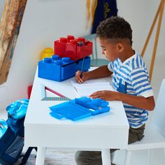 a young boy sitting at a white table writing on a piece of paper with legos in the background