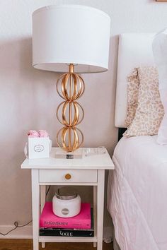 a nightstand with a lamp on top of it next to a pink book and white bed