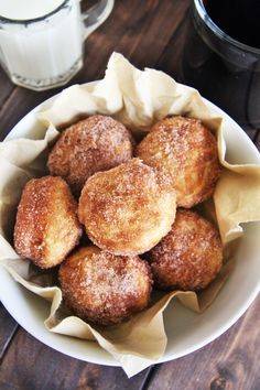 a white bowl filled with sugared donuts next to a glass of milk on top of a wooden table