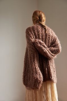 a woman standing in front of a white wall wearing a brown knitted sweater and beige pleated skirt