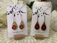 two red and black earrings are on display next to a white flower vase with flowers in it