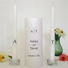 two white candles sitting on top of a table next to vases filled with flowers