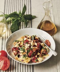 a white bowl filled with pasta and tomatoes next to a bottle of olive oil on a table