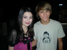 a boy and girl standing next to each other in front of a dark background with one smiling at the camera