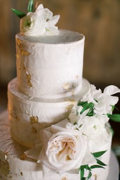 a three tiered cake with white flowers and greenery on the top is surrounded by gold flakes