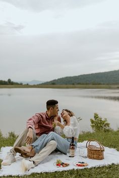 a man and woman are sitting on a blanket by the water eating food from bottles