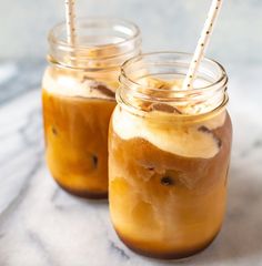 two mason jars filled with iced coffee sit on a marble counter top, one has straws in it