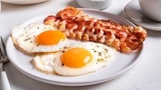 two eggs and bacon on a white plate with silverware next to coffee mugs