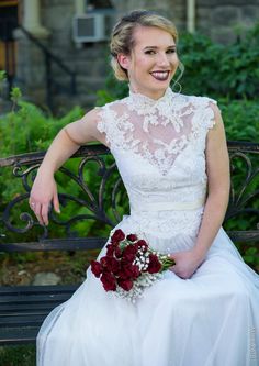 a woman sitting on a bench wearing a white dress and holding a red rose bouquet