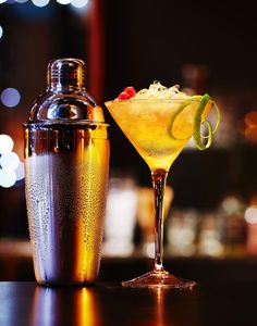 a drink in a glass next to a cocktail shaker on a table with blurry lights behind it