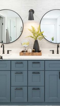 two round mirrors are above the double sinks in this modern bathroom with gray cabinets and white walls