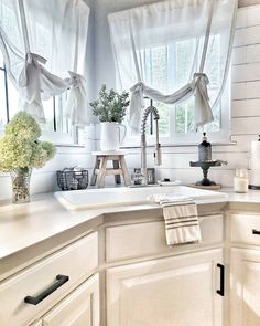 a kitchen with white cabinets and curtains on the window sill, along with flowers in vases