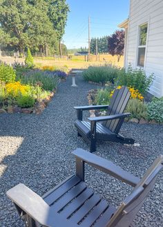 two chairs sitting on gravel in front of a white house and flower garden with yellow flowers