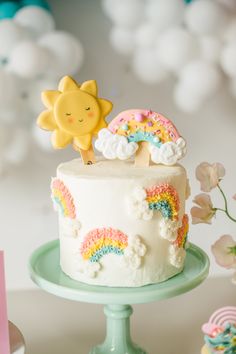 there is a white cake with rainbow decorations on the top, and a sun decoration above it