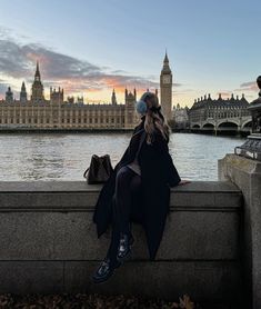 a woman is sitting on a ledge looking at the water and big ben in the background