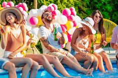 group of people sitting on the edge of a swimming pool with balloons in the background