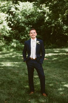 a man standing in the grass wearing a blue suit and white shirt with his hands in his pockets