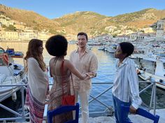 three women and one man are standing on a boat dock talking to each other, with boats in the water behind them