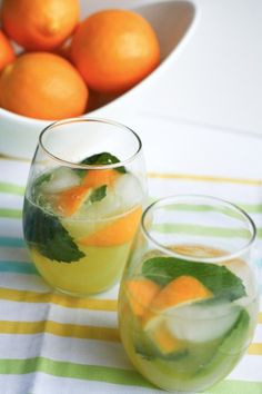 two glasses filled with lemonade and oranges next to a bowl of oranges