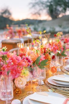 a long table is set with pink and orange flowers in glass vases, white plates and silverware