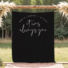 a black banner that says it's always you hanging from a wooden frame in front of some grass