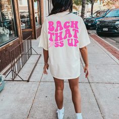 a woman walking down the sidewalk wearing a white shirt with pink letters on it that says back that s up