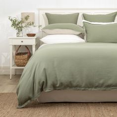 a bed with green sheets and pillows in a white room next to a basket full of plants