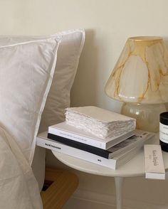 a white table topped with books and a lamp next to a pillow on top of a bed