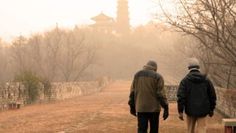 two people walking down a dirt road in the middle of winter with trees and buildings in the background