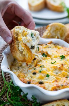 a person dipping some food into a white casserole dish with bread on the side