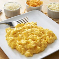 scrambled eggs on a square white plate with silver fork and small bowls of shredded cheese in the background