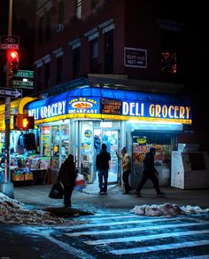 people walking in front of a deli grocery store on a city street at night