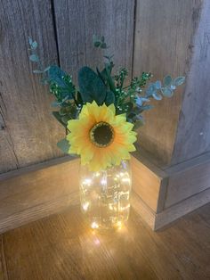 a sunflower in a mason jar with lights on the table next to a wooden wall