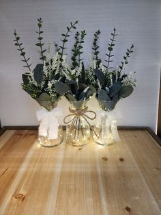 two glass vases with flowers and greenery in them on a wooden table top