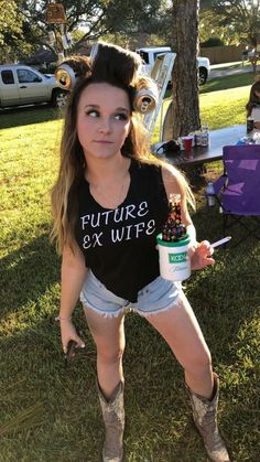 a woman in short shorts and boots holding a cup with dirt on her head while standing next to a tree