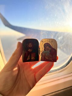 a hand holding two small wooden magnets depicting the virgin mary and jesus on an airplane window