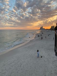 people walking on the beach at sunset with buildings in the background and clouds in the sky