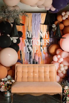 a room filled with balloons and an orange couch in front of a wall that says all my witches come