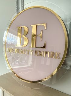 a sign that says be the beauty empire in gold lettering on a pink background with a mirror behind it