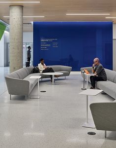 two people are sitting on couches in an office lobby with blue walls and white flooring