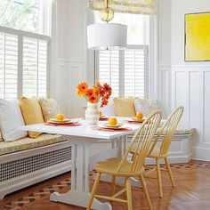 a white table with yellow chairs and plates on it in front of windows that have roman shades