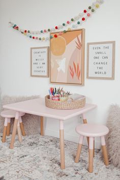 a pink table and two stools in front of a wall with pictures on it