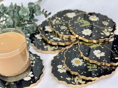 four black and gold coasters with daisies on them next to a cup of coffee