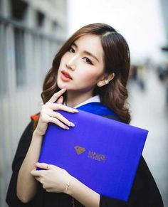 a woman in graduation gown holding a blue folder and looking at the camera with her finger on her lips