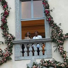 a man and woman standing on a balcony looking out the window at flowers growing all around them