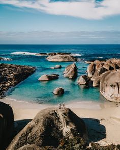 two people standing on the beach next to some large rocks and blue water with white clouds in the sky